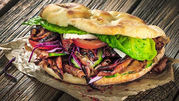 Ein gefülltes Fladenbrot mit gegrilltem Fleisch, Salat, Tomaten, Gurken, Rotkohl und Sauce auf braunem Papieruntergrund.
