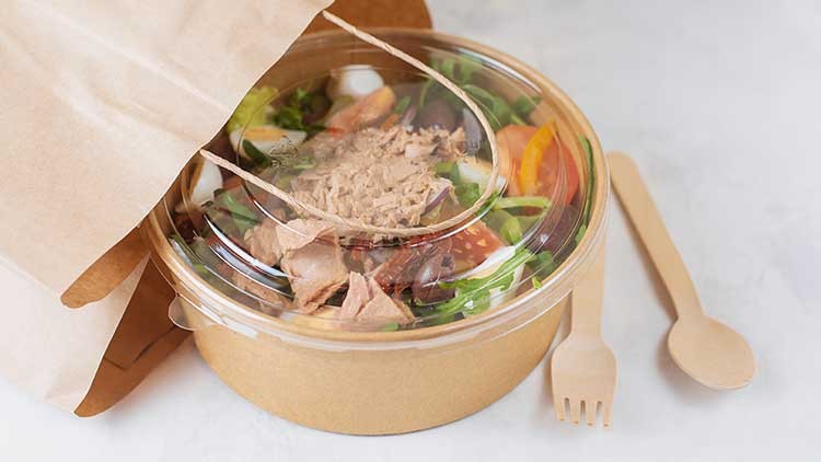 Ein braunes Pappbehältnis mit Salat und Thunfisch sowie durchsichtigem Deckel liegt neben Holzlöffel und -gabel.
