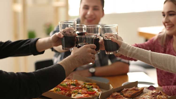 Vier Personen stoßen mit Gläsern dunkler Flüssigkeit über einem Tisch mit Pizza in Kartons an.