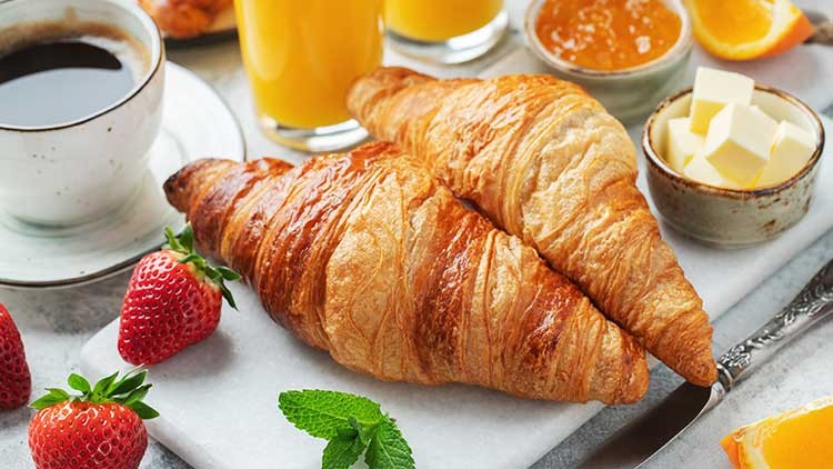 Zwei goldbraune Croissants liegen auf einem weißen Teller, umgeben von Erdbeeren, Orangenscheiben, Butter und Marmelade.