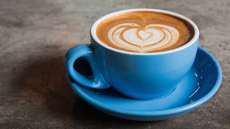 Eine blaue Tasse mit Cappuccino und Latte Art in Herzform steht auf einer passenden Untertasse auf einer grauen Tischplatte.