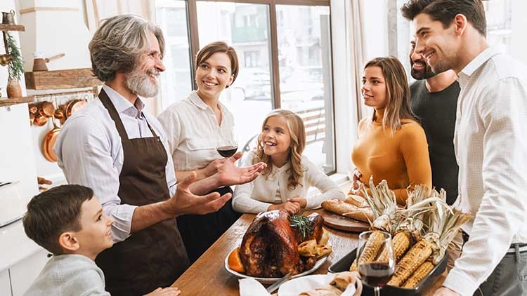 Mehrere Personen stehen an einem gedeckten Holztisch mit gebratener Pute, Maiskolben, Brot und Gläsern mit Rotwein.