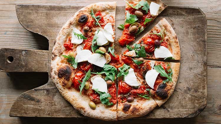 Knusprige Pizza mit roten Tomaten, grünem Rucola, Oliven und weißem Mozzarella, serviert auf einem rustikalen Holzbrett.