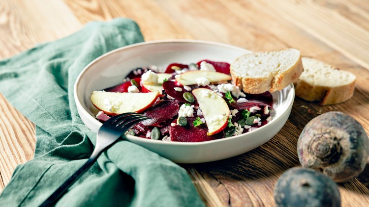 Weiße Schale mit Rote-Bete-Salat, Apfelscheiben, Kürbiskernen, Käse, Brot und schwarzer Gabel auf grünem Tuch und Holzuntergrund.