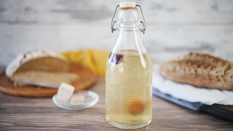 Eine Glasflasche mit durchsichtigem Getränk steht vor zwei Brotscheiben und einem kleinen Teller mit Hefewürfeln.