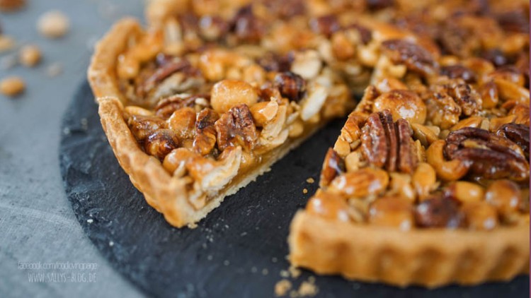 Runde Tarte mit karamellisierten Nüssen wie Pekannüssen und Mandeln, auf einer schwarzen Schieferplatte, ein Stück fehlt.