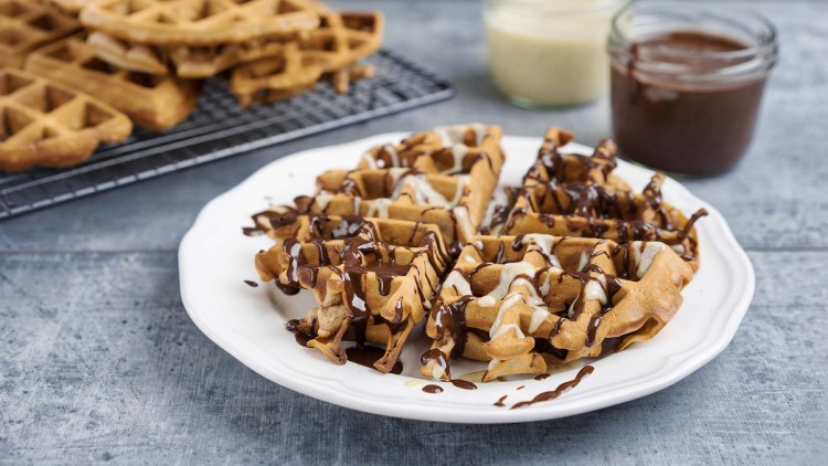 Ein Teller mit vier Waffelstücken, dekoriert mit heller und dunkler Soße, daneben Glasbehälter mit Schokosoße und Vanillesoße.