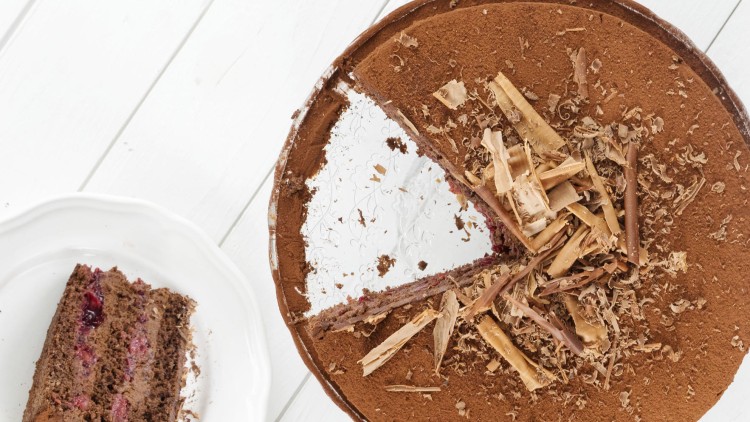 Runde Schokoladentorte mit Schokoraspeln, ein Stück fehlt und liegt auf einem weißen Teller, gefüllt mit Kirschen und Creme.