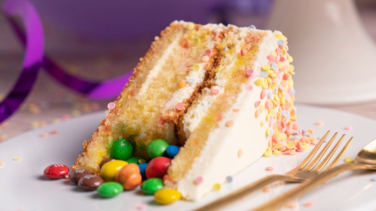 Ein Stück heller Schichttorte mit Zuckerdekor und bunten Schokolinsen liegt auf einem Teller neben goldfarbenem Besteck.
