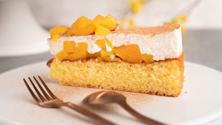 Ein Stück Kuchen mit gelbem Biskuitboden, Sahneschicht, Pfirsichstücken und Zimt liegt auf einem weißen Teller mit Besteck.