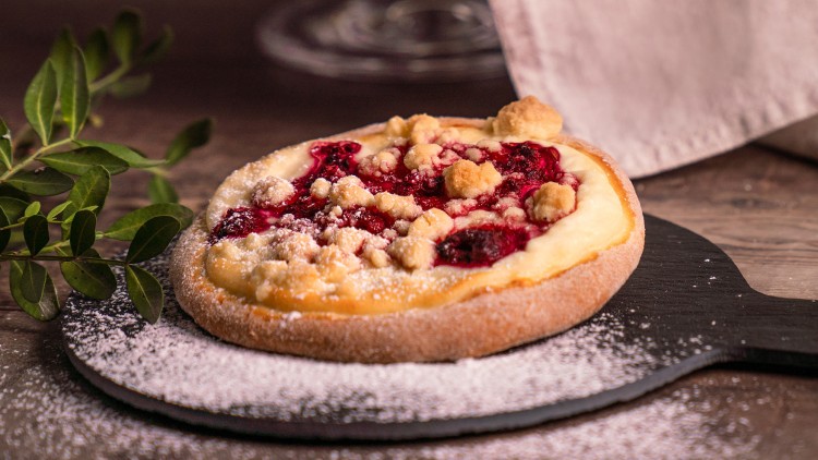 Kleiner Obstkuchen mit Streuseln, Puderzucker und roten Fruchtfüllungen auf schwarzer Holzunterlage, daneben grüne Blätter.