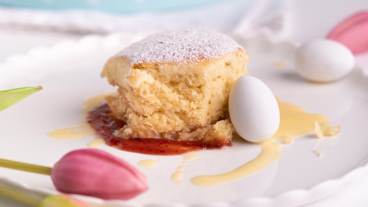 Ein Stück heller Kuchen mit Puderzucker, Marmelade und heller Soße auf einem Teller, dekoriert mit zwei weißen Schokoeiern und Tulpen.