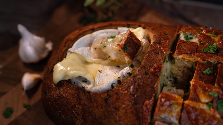 Ein Laib Brot mit Schlitzen, gefüllt mit geschmolzenem Käse und Kräutern, daneben Knoblauchzehen auf einem Holzbrett.