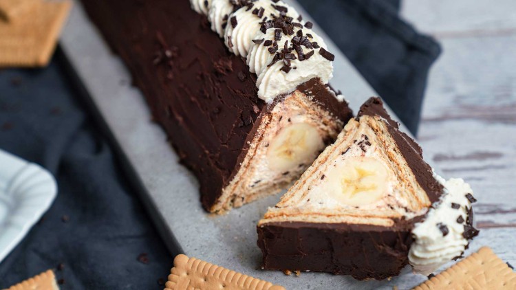 Dreieckiger Schokoladenkuchen mit Bananenfüllung, Keksboden und Sahnedekoration, auf einem grauen Untergrund angeschnitten.
