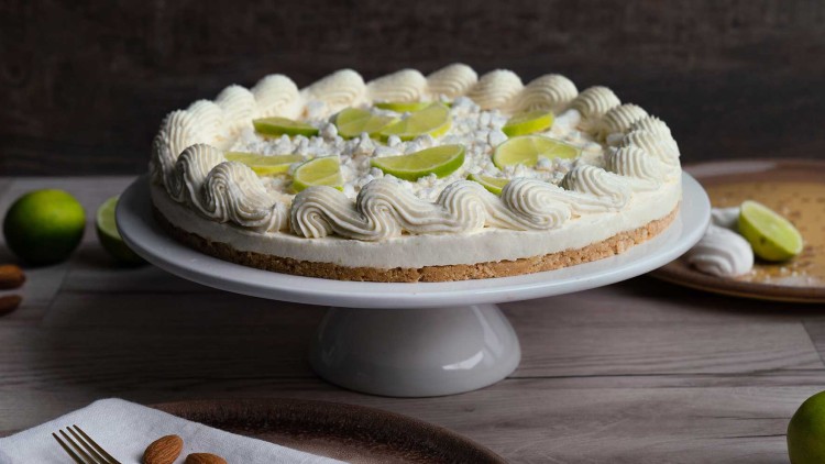 Eine helle Torte mit Sahne, Limettenscheiben und Krümeln auf einem weißen Tortenteller aus Keramik, dekoriert mit Mandeln.