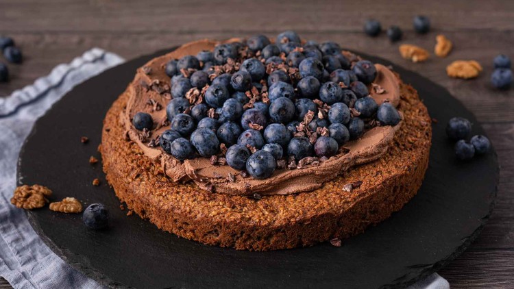Runder Schokoladenkuchen mit Schokocreme, frischen Blaubeeren und Kakaonibs auf einer dunklen Schieferplatte.