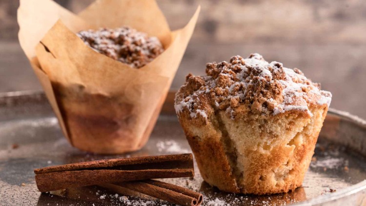 Zwei Muffins mit Streuseln und Puderzucker auf einem Blech, davor liegen zwei Zimtstangen.