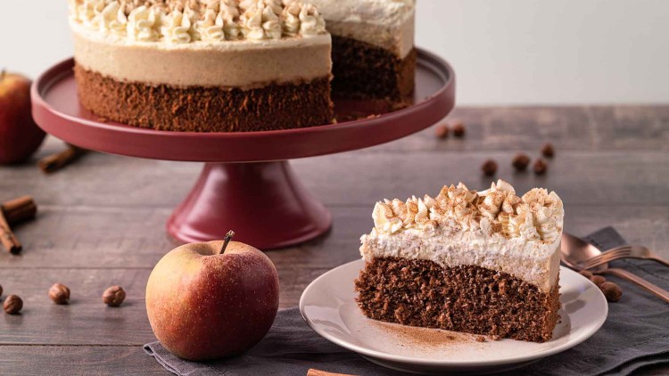 Schichttorte mit Sahnehäubchen und Zimt auf einem roten Tortenständer, daneben ein Apfel und ein angeschnittenes Stück auf einem Teller.