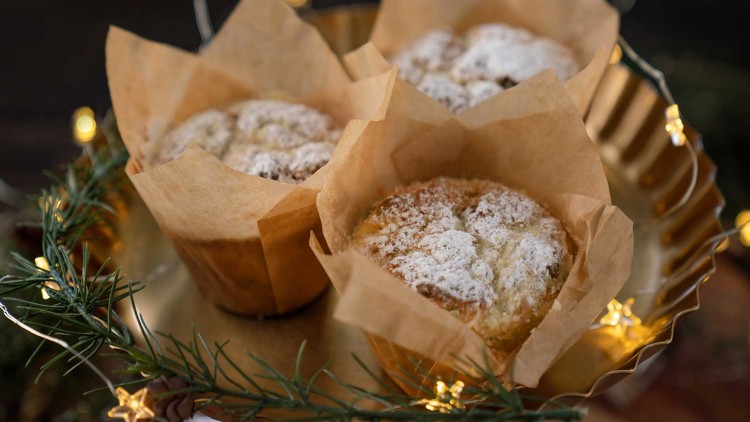 Drei Muffins in braunem Backpapier mit Puderzucker auf einer goldenen Platte, verziert mit Tannenzweigen und Lichterketten.