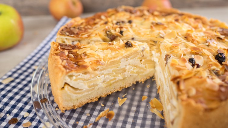 Aufgeschnittene Apfeltorte mit Mandelsplittern und Rosinen auf kariertem Tuch, im Hintergrund Äpfel auf unscharfem Untergrund.