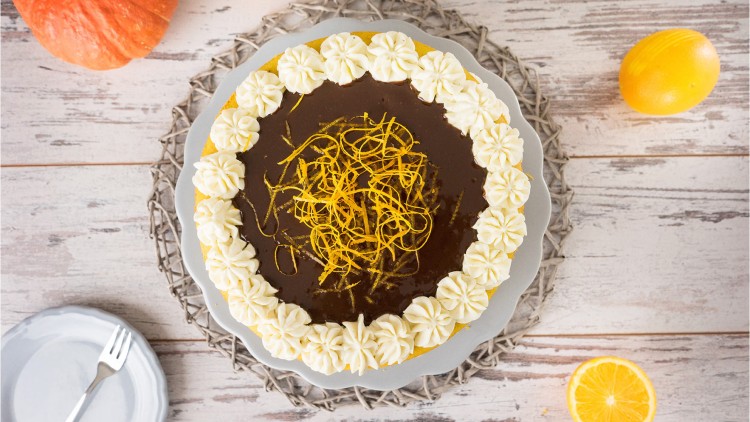 Runder Kuchen mit Schokoladenglasur, Sahnetuffs am Rand und Zesten aus Orangenschale auf einem Teller, daneben Kürbis und Orange.