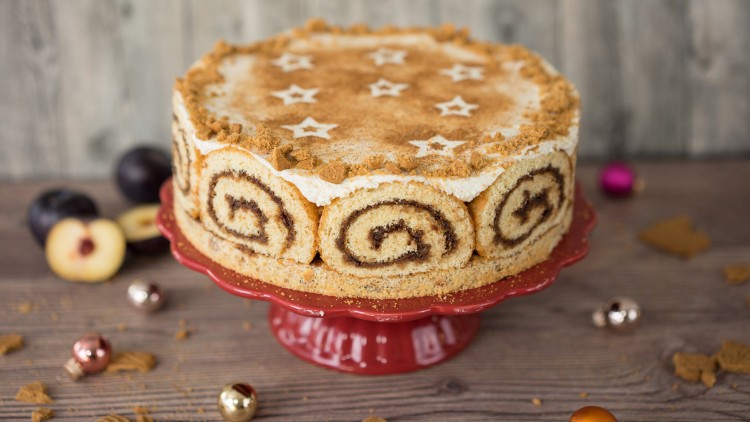Runde Torte aus Biskuitrollen mit Sahne, Zimt und Sternen aus Puderzucker auf roter Tortenplatte, Dekoration mit Zwetschgen.