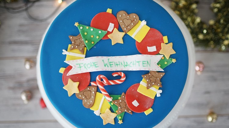 Runder Kuchen mit blauer Glasur, weihnachtlicher Zuckerdekoration und Schriftzug Frohe Weihnachten auf einem weißen Banner.