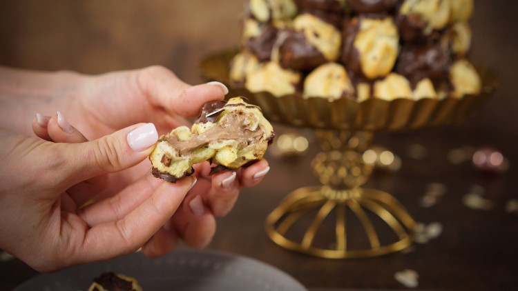 Zwei Hände halten ein aufgebrochenes, mit Schokoladencreme gefülltes Gebäck, im Hintergrund ein goldener Servierteller mit ähnlichen Gebäcken.