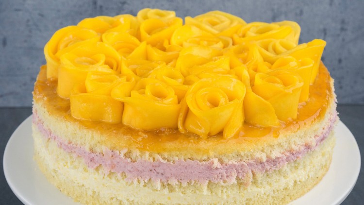 Runde Torte mit gelben Mangostreifen in Rosenform auf der Oberfläche und heller Cremefüllung mit rosa Schicht auf weißem Teller.