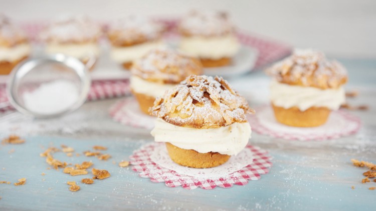 Kleine Gebäckstücke mit heller Cremefüllung und knuspriger Zuckerkruste, dekoriert mit Puderzucker auf kleinen Spitzendeckchen.