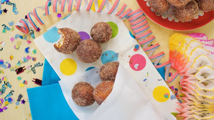 Braune, zuckerbestreute Teigbällchen liegen auf buntem Punktetuch, umgeben von Konfetti, Papierschlangen und Dekoration.