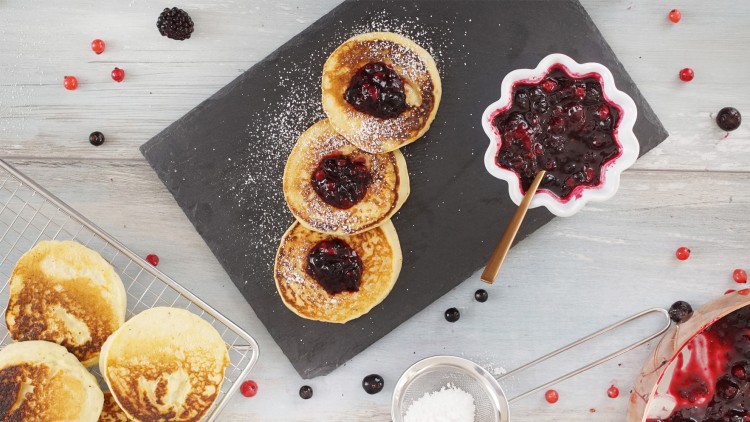 Drei Pancakes mit roter Beerenmarmelade und Puderzucker liegen auf schwarzem Schiefer, daneben Sieb und Beeren in Schalen.