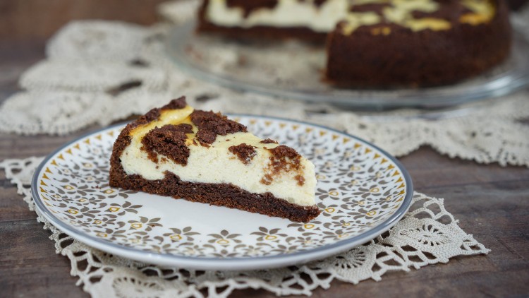 Ein Stück russischer Zupfkuchen liegt auf einem Teller mit floralem Muster, dahinter eine Kuchenplatte auf einem Spitzendeckchen.