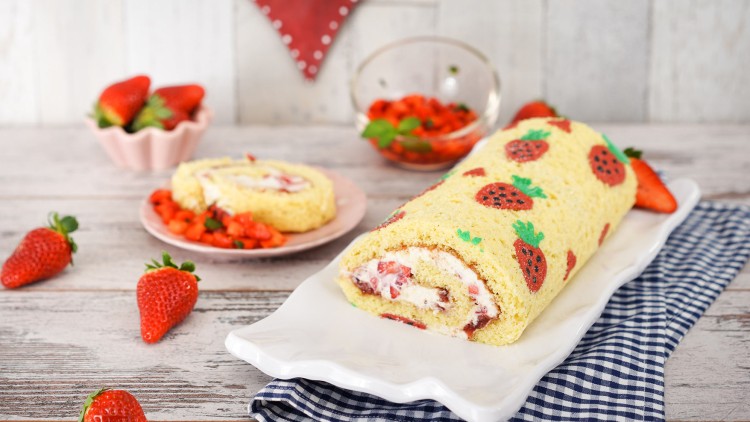 Biskuitrolle mit Erdbeer-Muster und Sahnefüllung liegt auf einer weißen Platte, umgeben von frischen Erdbeeren und Deko.