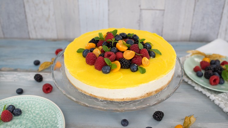 Käsekuchen mit gelber Fruchtschicht, dekoriert mit Himbeeren, Blaubeeren, Brombeeren, Physalis und Minzblättern auf Glasplatte.