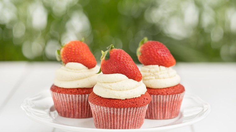 Drei rote Cupcakes mit weißem Sahnetopping und je einer Erdbeere darauf stehen auf einem weißen Teller.