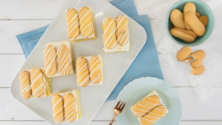 Rechteckige Kuchenstücke mit hellem Boden, Sahneschicht und Löffelbiskuits mit Zuckerglasur liegen auf einer weißen Platte.