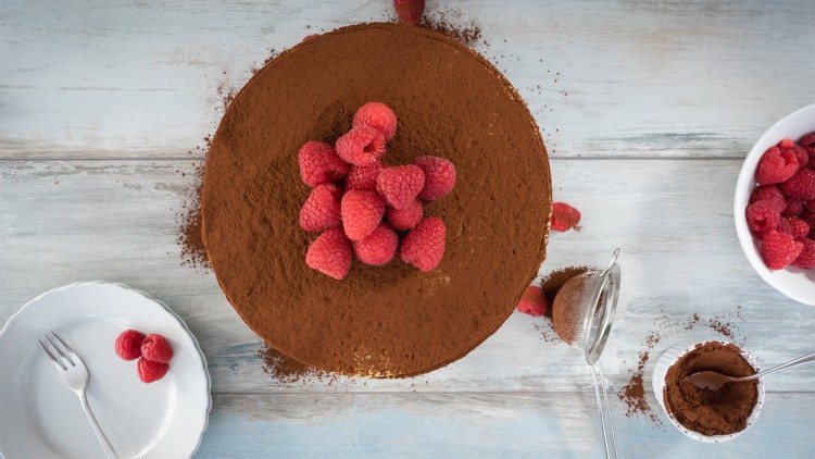 Runde Torte mit Kakaopulver bestäubt, darauf frische Himbeeren, daneben Teller, Sieb, Schale mit Himbeeren und Kakaopulver.