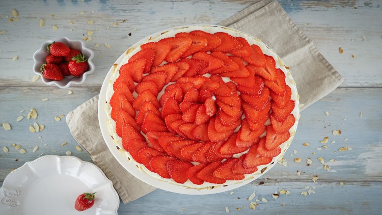 Erdbeerkuchen mit spiralförmig angeordneten Erdbeerscheiben auf weißem Teller, daneben Schale mit Erdbeeren und leeres Teller.