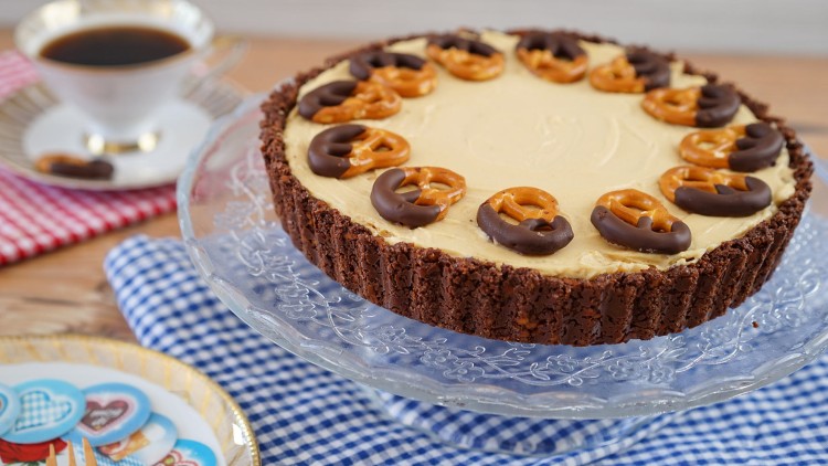 Runder Kuchen mit heller Creme und Schoko-Brezeln auf einem Glasständer, im Hintergrund Tasse Kaffee und bunte Servietten.