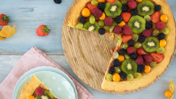 Runder Obstkuchen mit Kiwi, Brombeeren, Himbeeren und Trauben auf Holzplatte, ein Kuchenstück auf einem gepunkteten Teller.