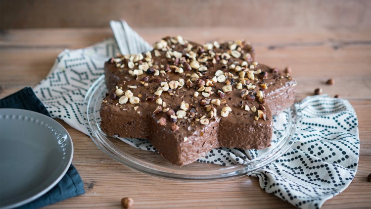 Schokokuchen in Sternform mit gehackten Haselnüssen auf Glasplatte, darunter gemustertes Tuch, daneben grauer Teller.