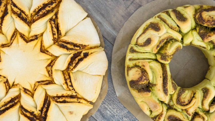 Zwei runde Brote auf Backpapier, links ein sternförmiges Brot, rechts ein grünes gefülltes Kranzbrot mit sichtbaren Kräutern.
