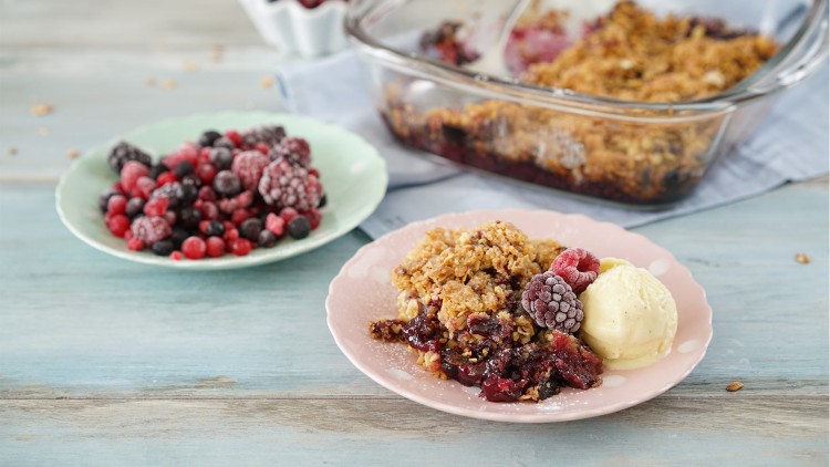 Ein Teller mit Beeren-Crumble und Vanilleeis steht vor einer Glasschale mit Crumble und einem Teller gefrorener Beeren.