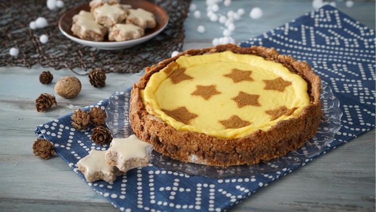Runder Kuchen mit gelber Füllung und braunen Sternen aus Zimt, Zimtsterne und Deko aus Tannenzapfen auf blauem Tuch.