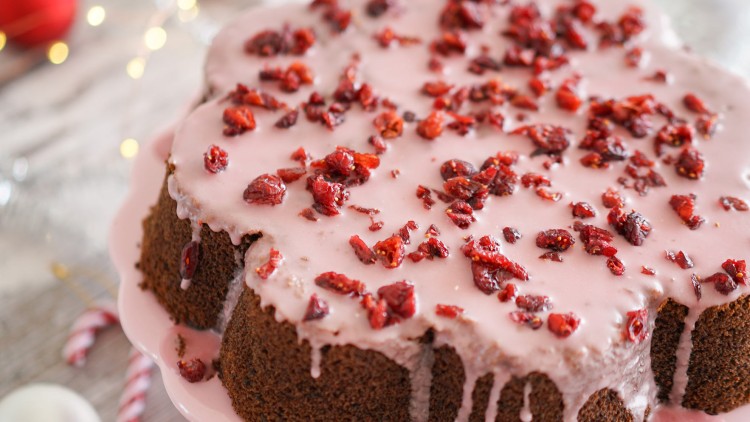 Ein Gugelhupf mit rosa Zuckerguss und getrockneten roten Beeren auf einer rosa Platte, im Hintergrund Weihnachtsdeko.