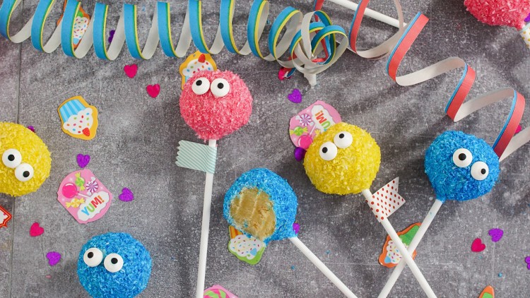 Bunte Cake Pops mit Augen und Kokosraspeln liegen auf grauem Untergrund, dekoriert mit Party-Streudeko und Luftschlangen.