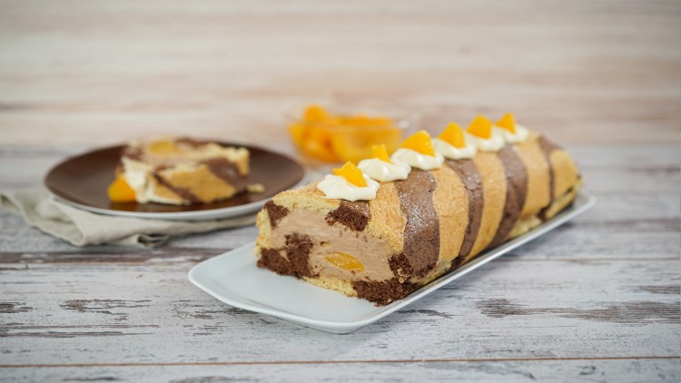 Biskuitrolle mit braunen und hellen Streifen gefüllt mit Creme und Pfirsichstücken, dekoriert mit Sahnehäubchen und Pfirsichen.