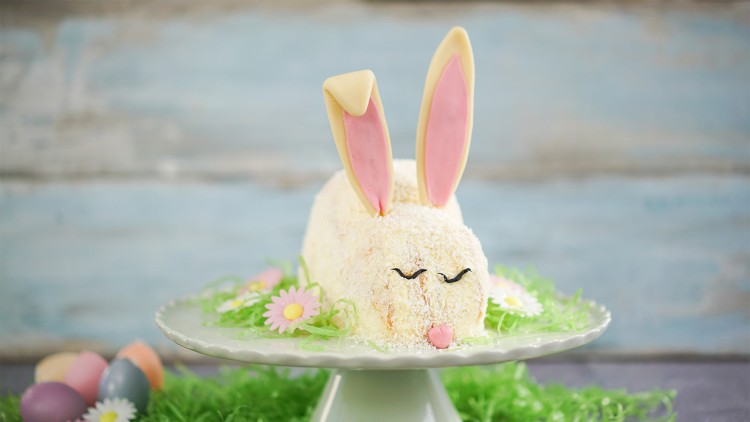 Kuchen in Form eines weißen Hasen mit rosa Ohren auf einer weißen Tortenplatte, dekoriert mit grünem Gras und Blüten.