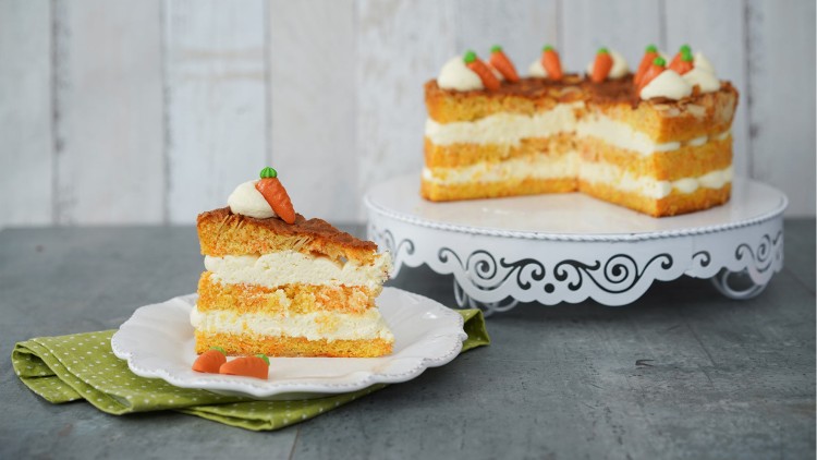 Ein Stück Karottentorte mit Frischkäsecreme liegt auf einem Teller, daneben steht die Torte auf einer weißen Tortenplatte.
